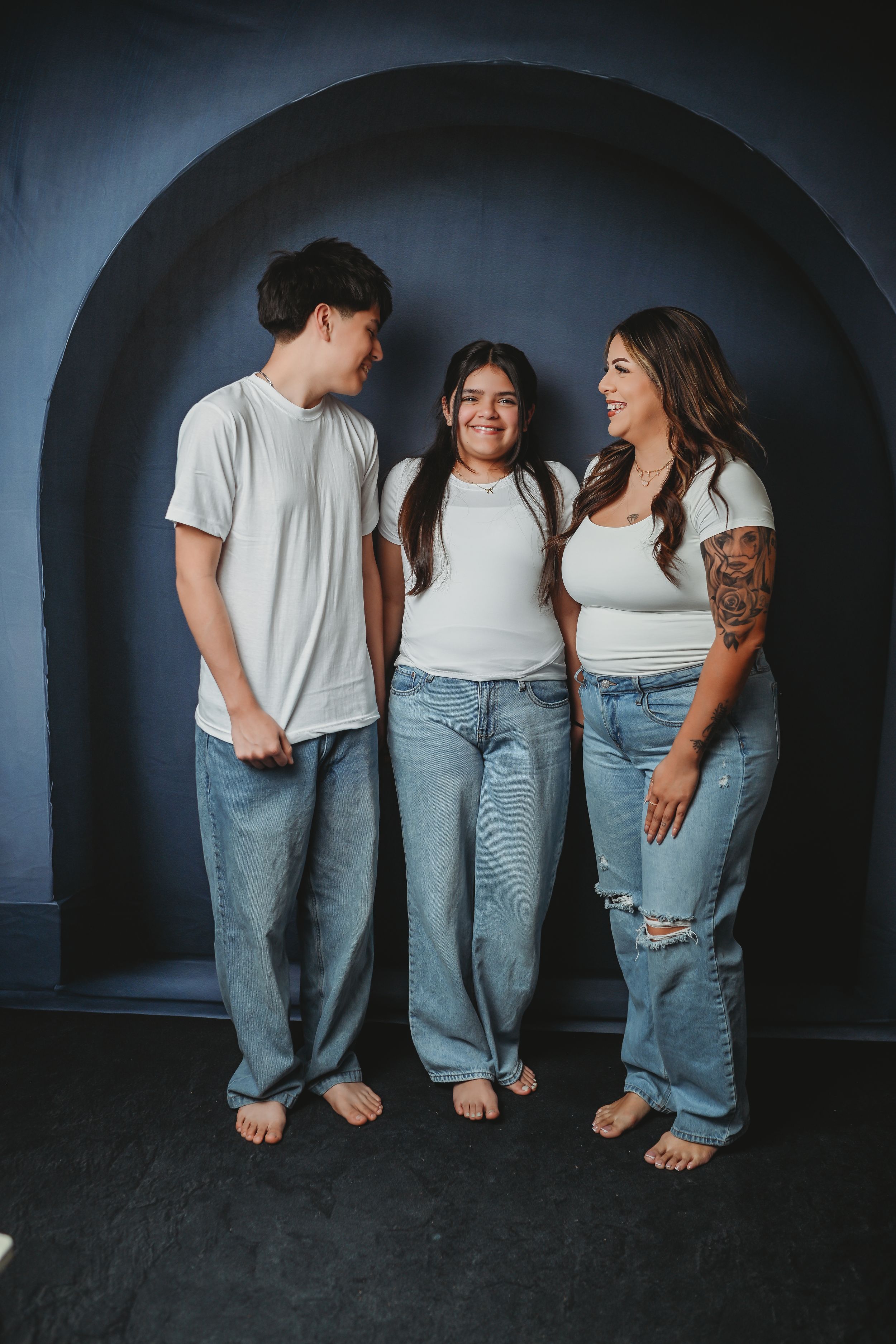 Three barefoot people in white tees and jeans smile together against a dark arched wall backdrop.
