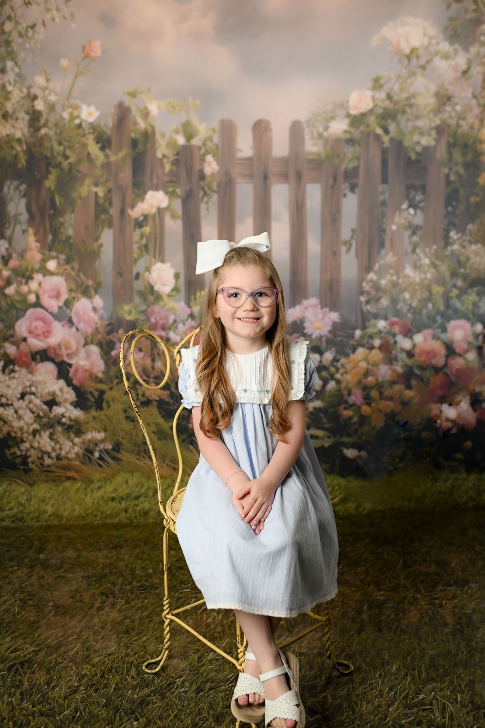 A smiling young girl in a blue dress and glasses sits on a gold chair before a floral garden backdrop.