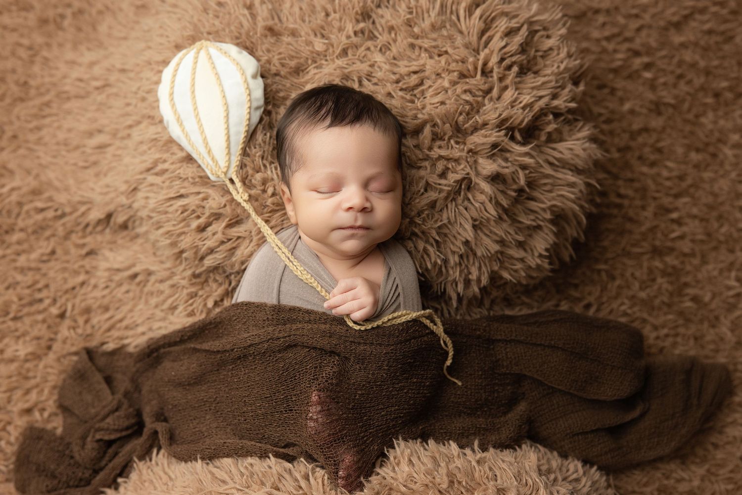 Newborn baby wrapped in teal swaddle holding green knit bear toy, resting in decorative bucket.