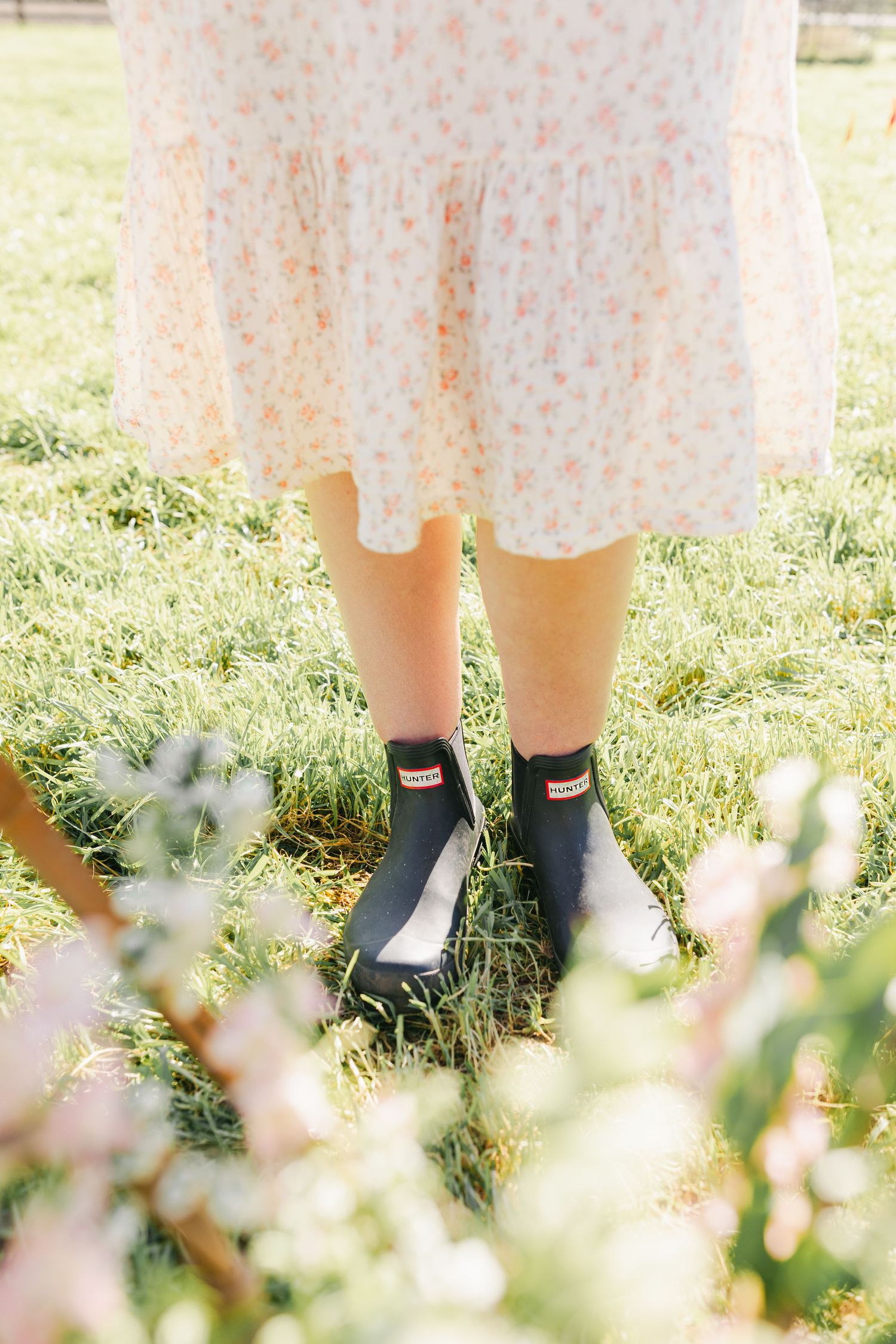 Woman in floral dress and black Hunter chelsea boots standing on green grass with pink flowers in foreground.