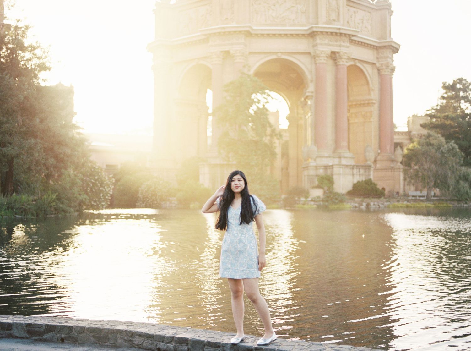 Young woman in white dress standing by a reflective pond with a grand arched monument at golden hour.