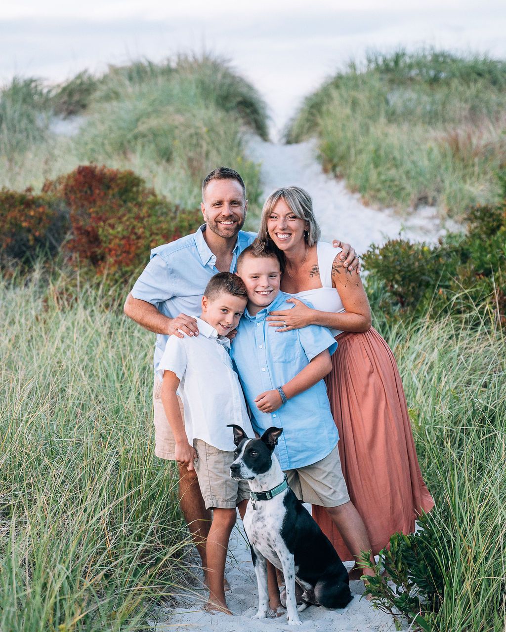 Happy family of four with a dog posing on a sandy beach path surrounded by dune grass.