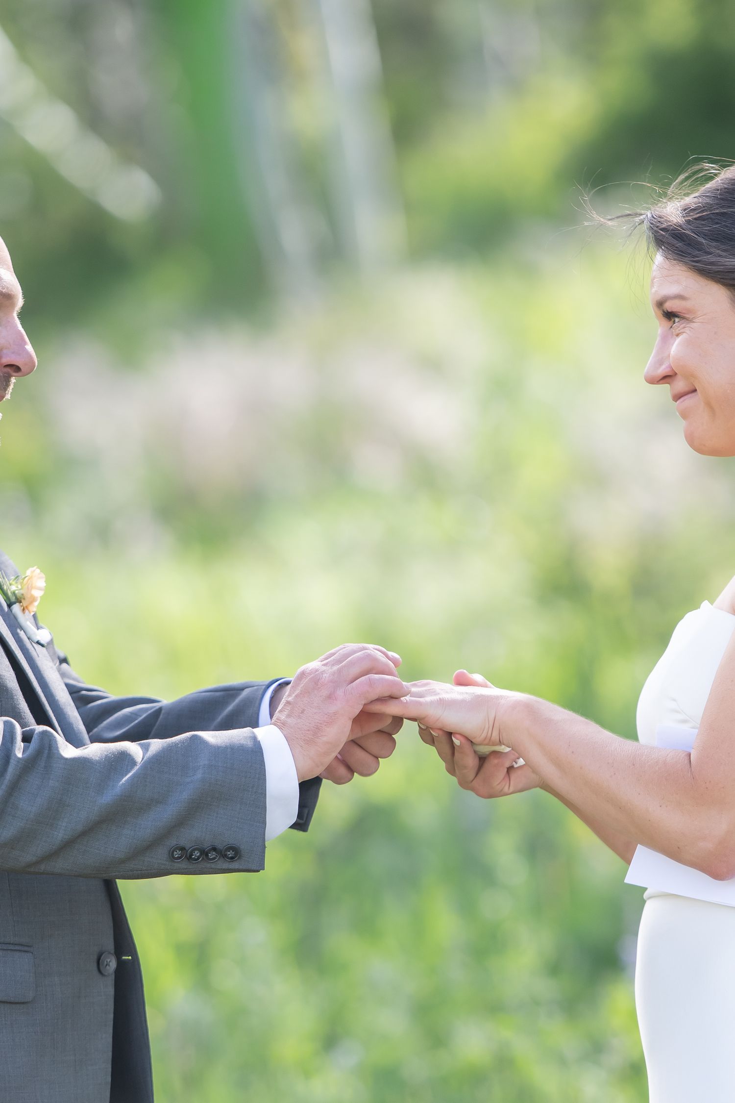 Bride and groom exchanging wedding rings outdoors during a sunny ceremony with green nature background.