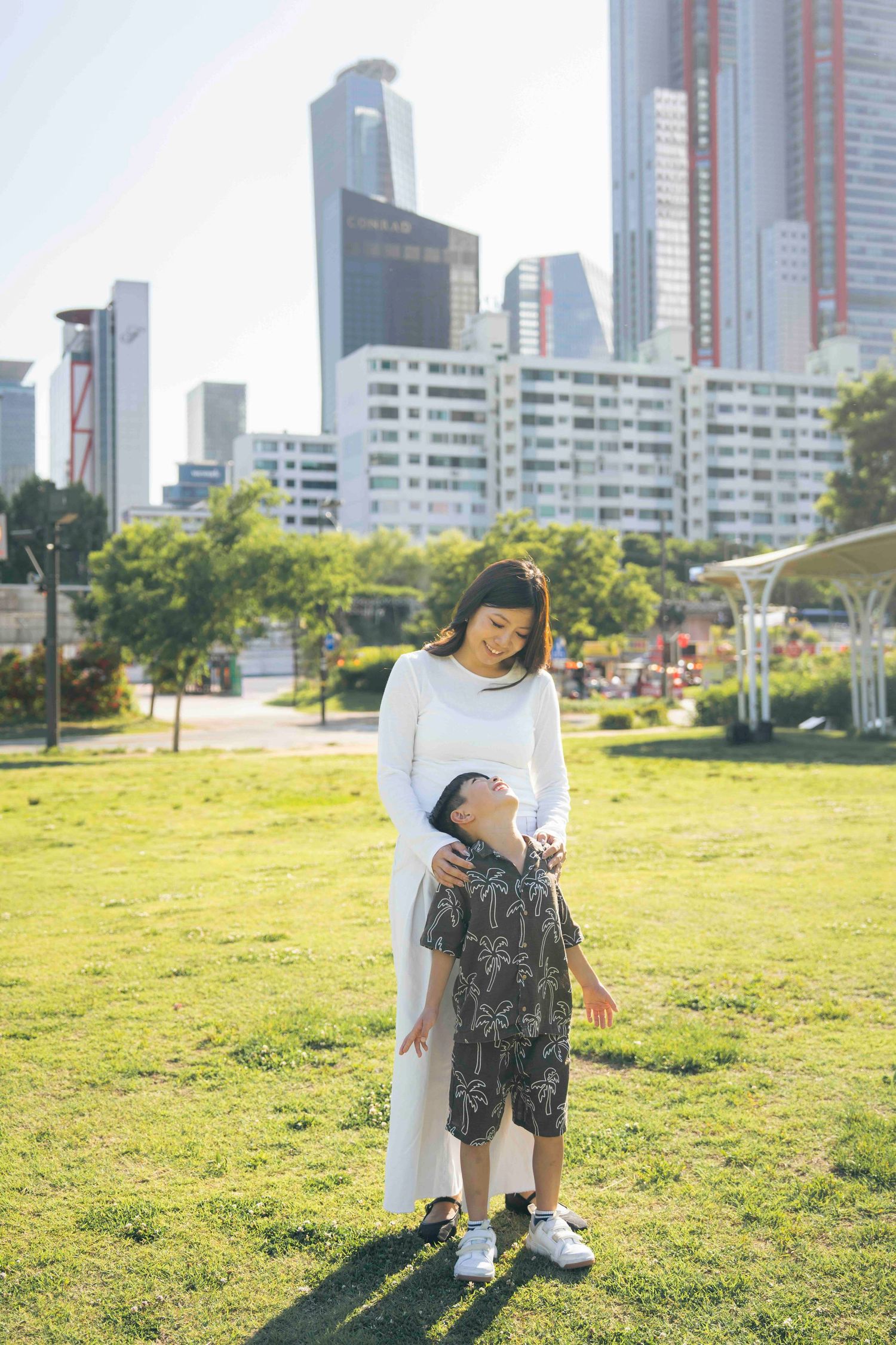 A mother in white dress embraces her young son in a sunny urban park with modern skyscrapers in the background.