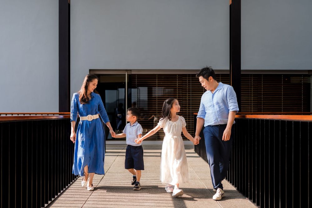 Happy Asian family of four holding hands walking together outside a modern building on a sunny day.