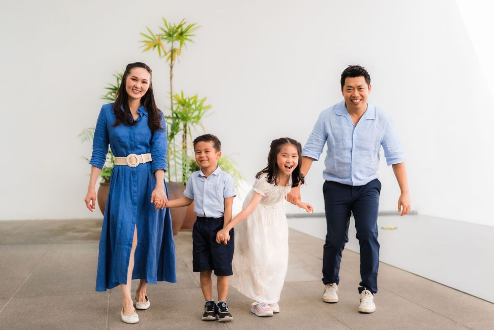 Happy Asian family of four posing together, parents and two young children smiling in a bright, modern white space.