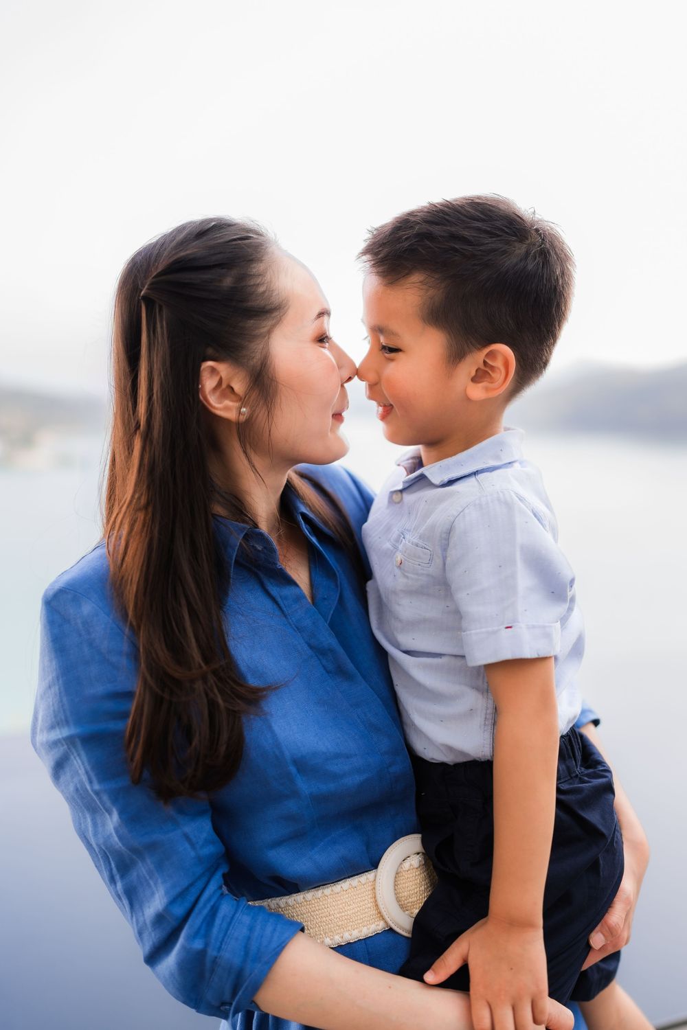 A mother in a blue dress holds her young son close as they touch noses and smile at each other outdoors.