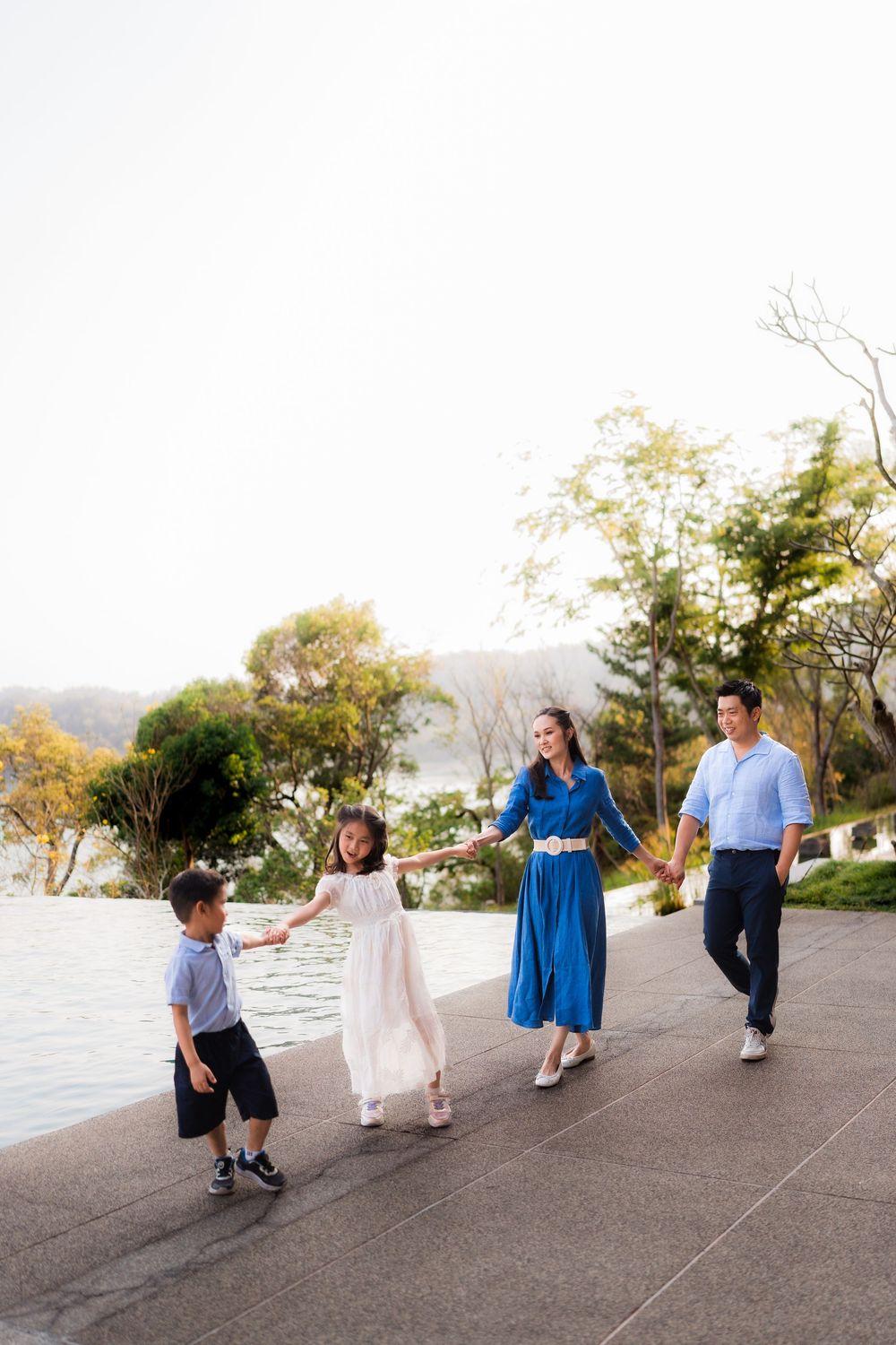 A happy Asian family of four walks hand-in-hand along a scenic lakeside path surrounded by trees.