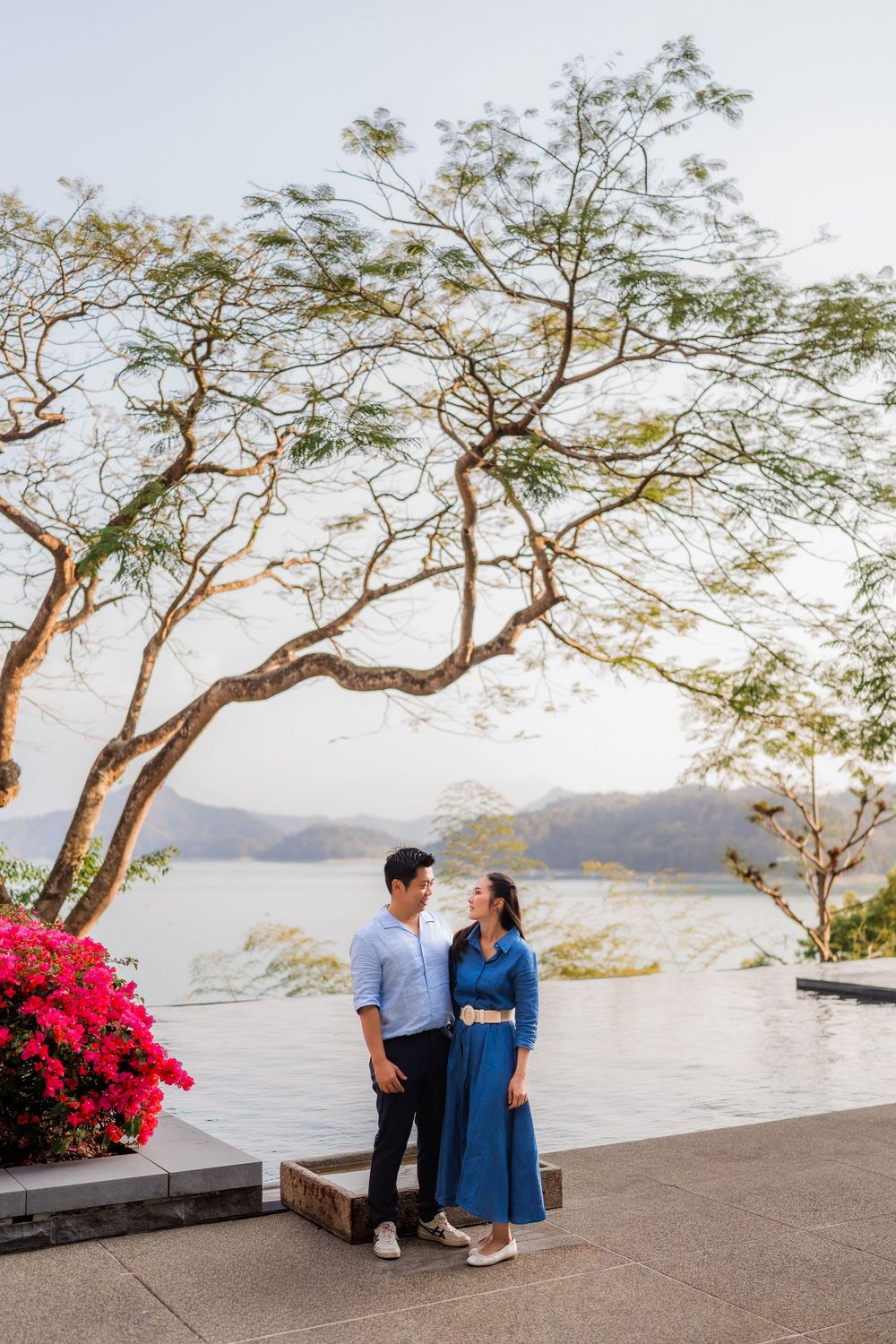 Couple standing together by a serene lake with pink flowers and trees at golden hour.