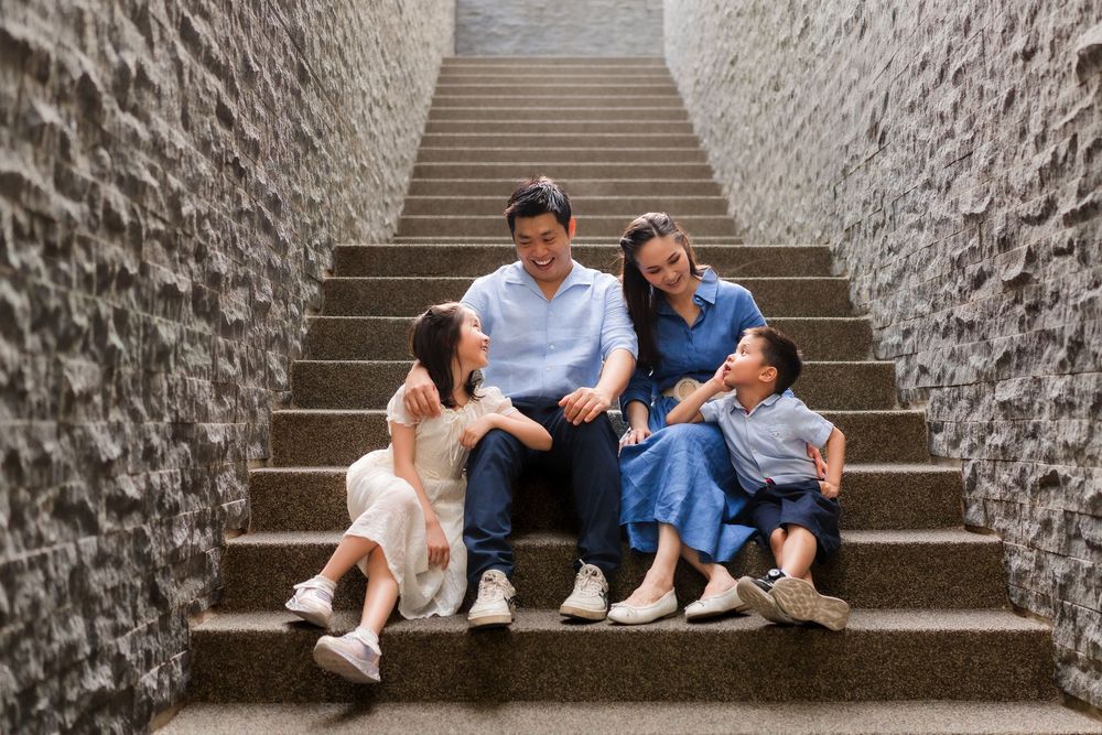 A happy Asian family of four sits together on stone steps between textured walls, sharing a joyful moment.