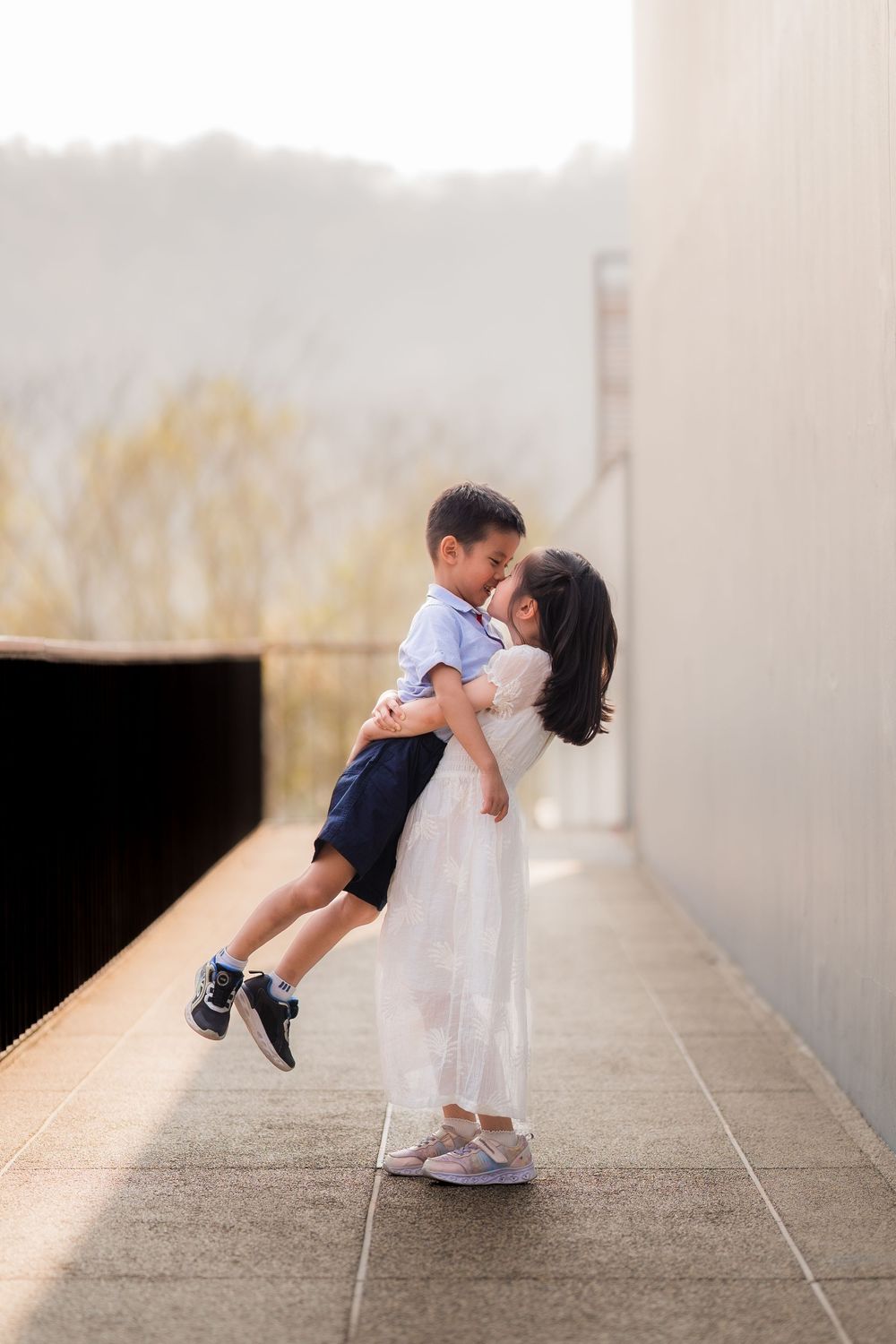 A girl in a white dress lifts and hugs a young boy on a tiled outdoor corridor with misty trees in the background.