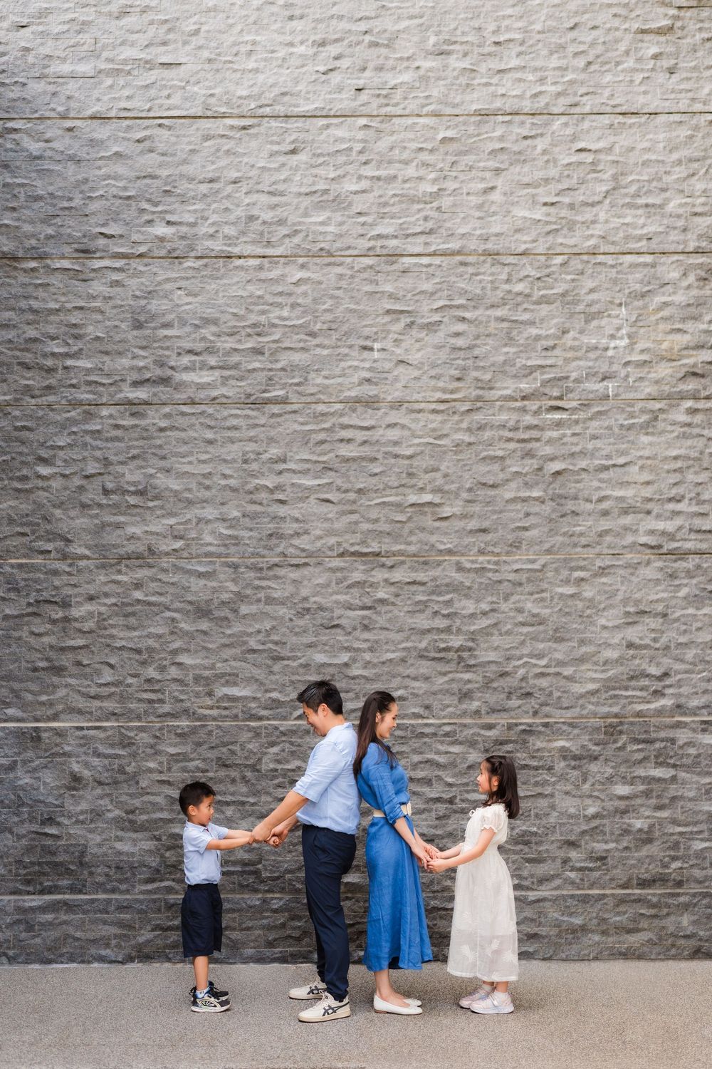 Family of four holding hands in front of a gray stone wall, parents in blue outfits with two young children.