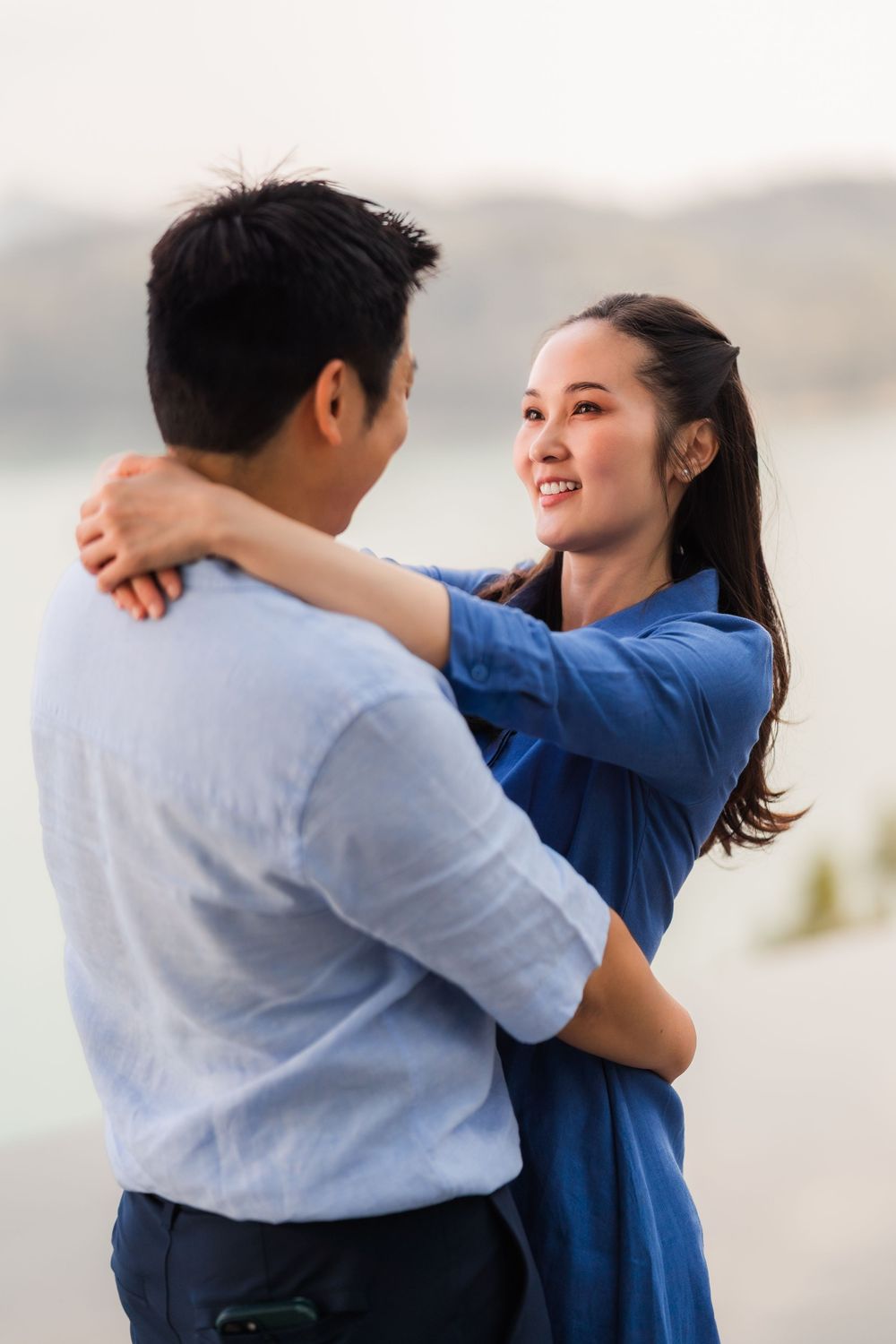 Smiling Asian woman embracing man outdoors near a calm lake at dusk.