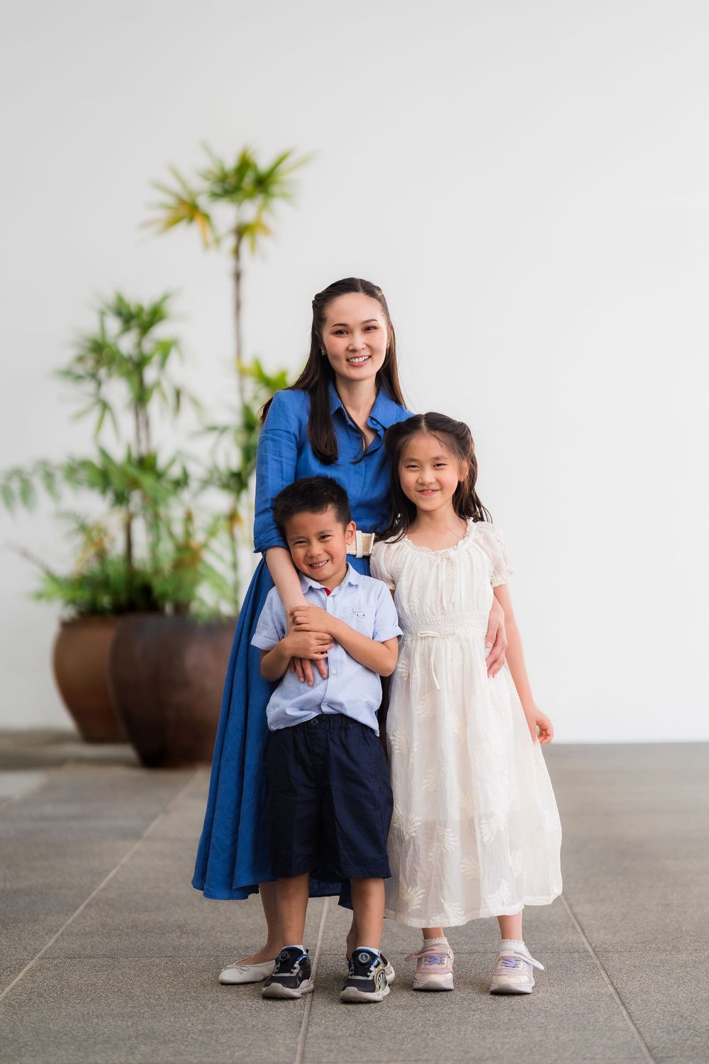 A smiling Asian mother in a blue dress hugs her two young children, a boy and girl, outdoors against a white wall.