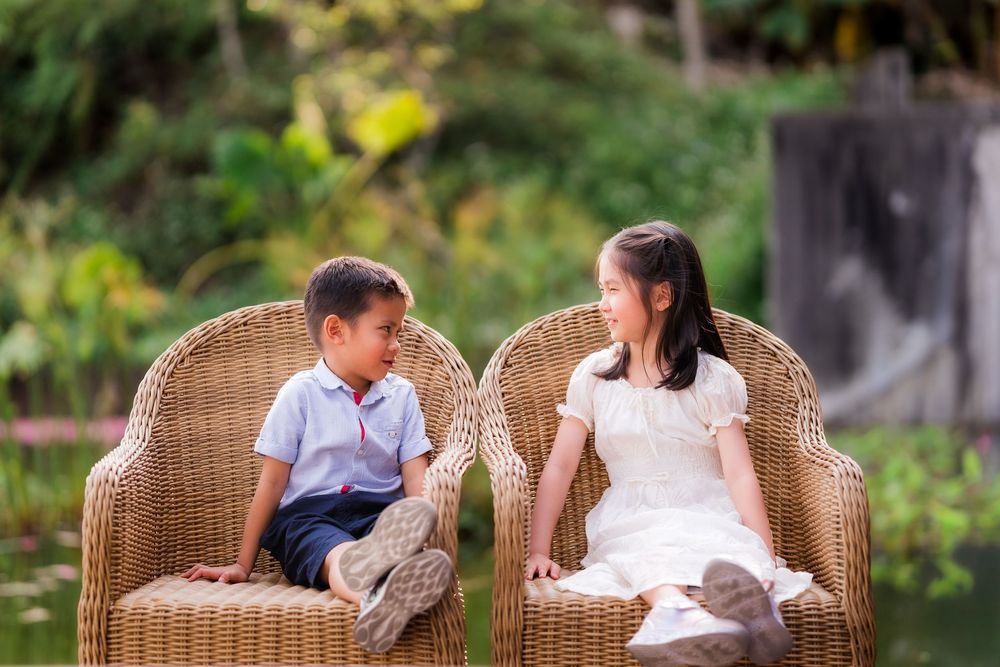 Two young children smile at each other while sitting in wicker chairs outdoors in a lush green garden.