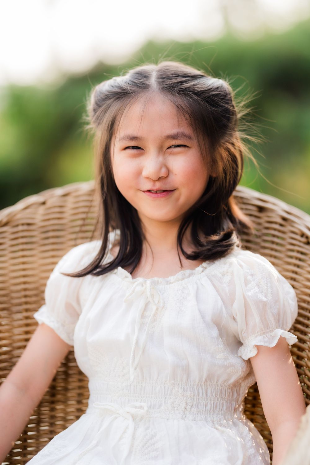 Cute Asian girl in white dress sitting in wicker chair, smiling outdoors in natural light.