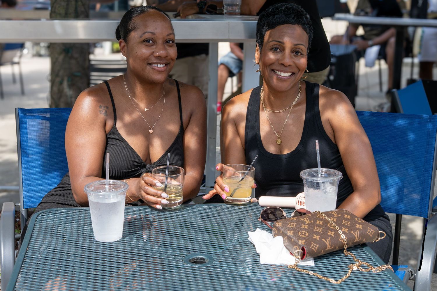 Two women smiling at outdoor cafe table with drinks and Louis Vuitton bag on sunny day.