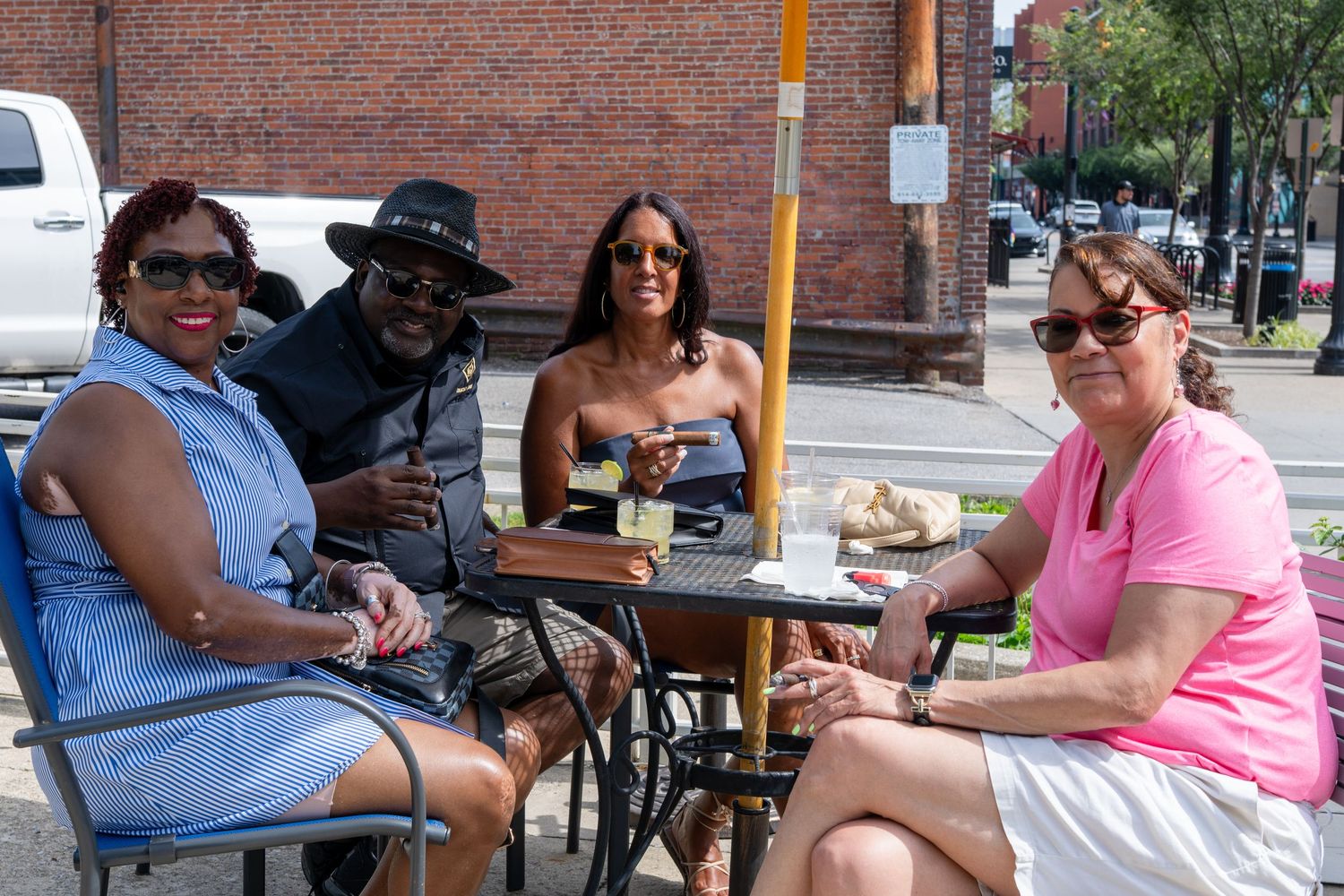 Four friends smile at an outdoor cafe table on a sunny day, enjoying drinks near a brick building.