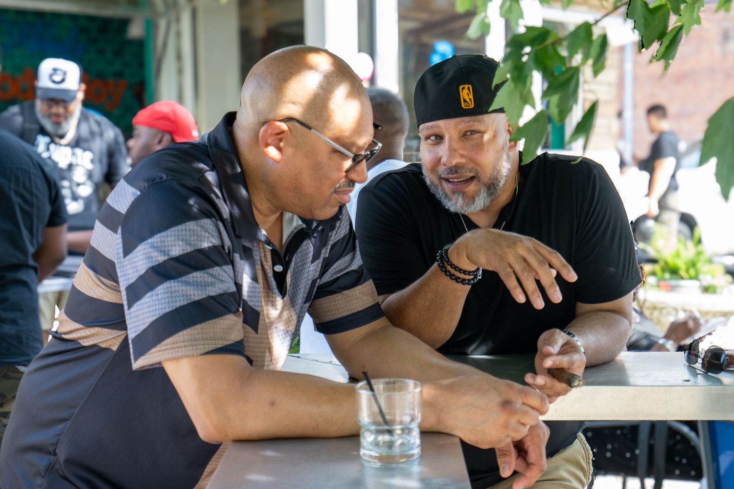 Two men in caps lean close in conversation at an outdoor table, with a crowd gathered in the background.