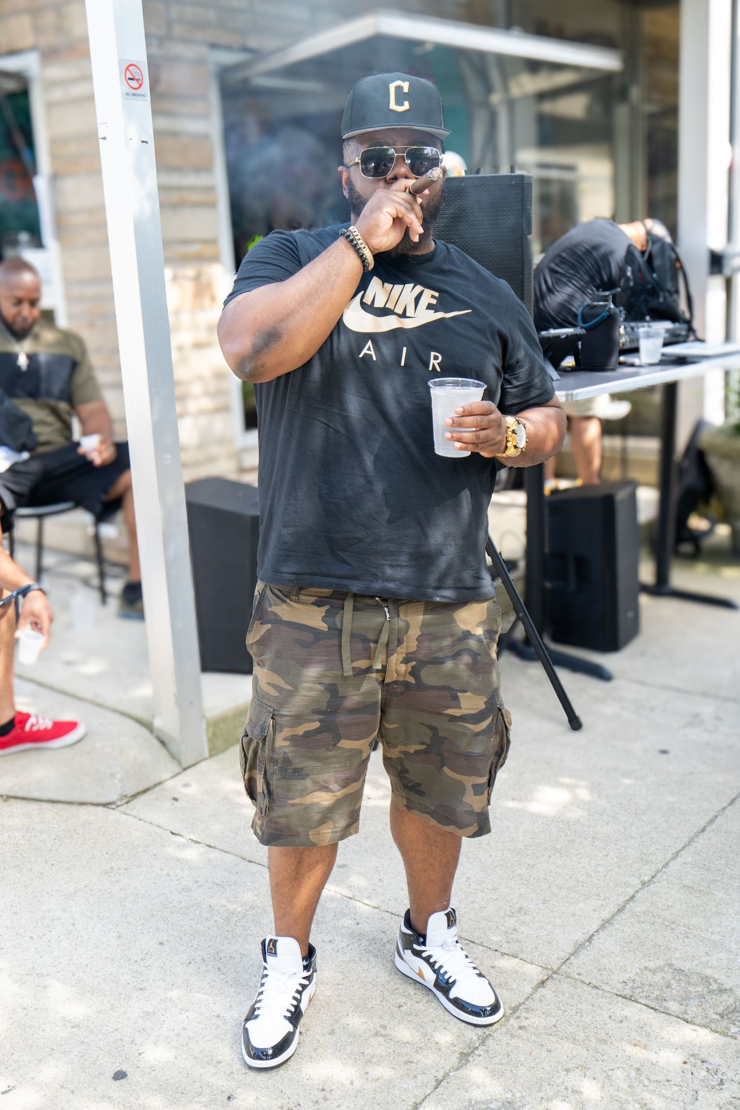 Man in Nike Air shirt, camo shorts, and Jordan sneakers holding a cup at an outdoor event.