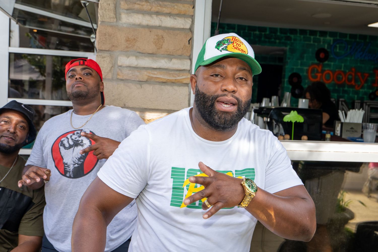 Two men pose outdoors near a food stand, wearing casual streetwear including caps and graphic t-shirts.