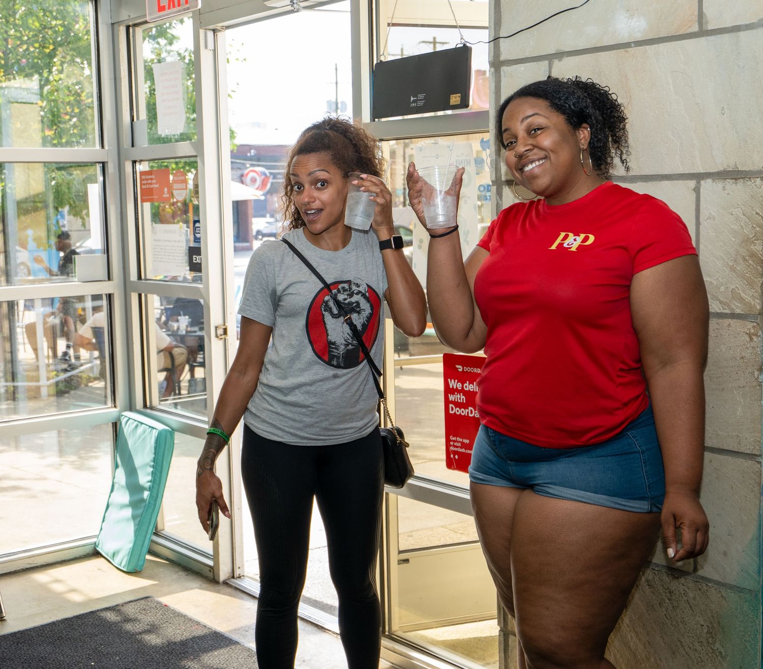 Two women smiling and chatting near a store entrance, one wearing a red P&P shirt and denim shorts.