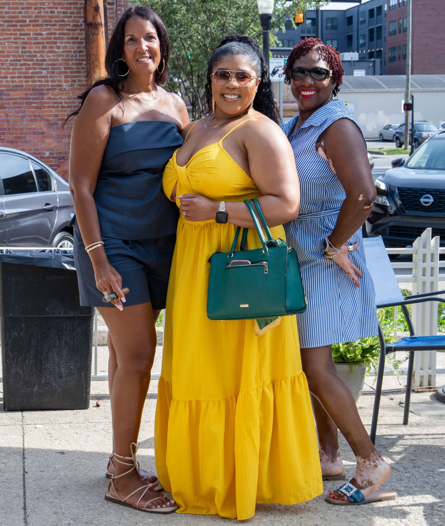 Three women posing outdoors, wearing colorful summer outfits in blue, yellow, and striped dresses on a sunny day.