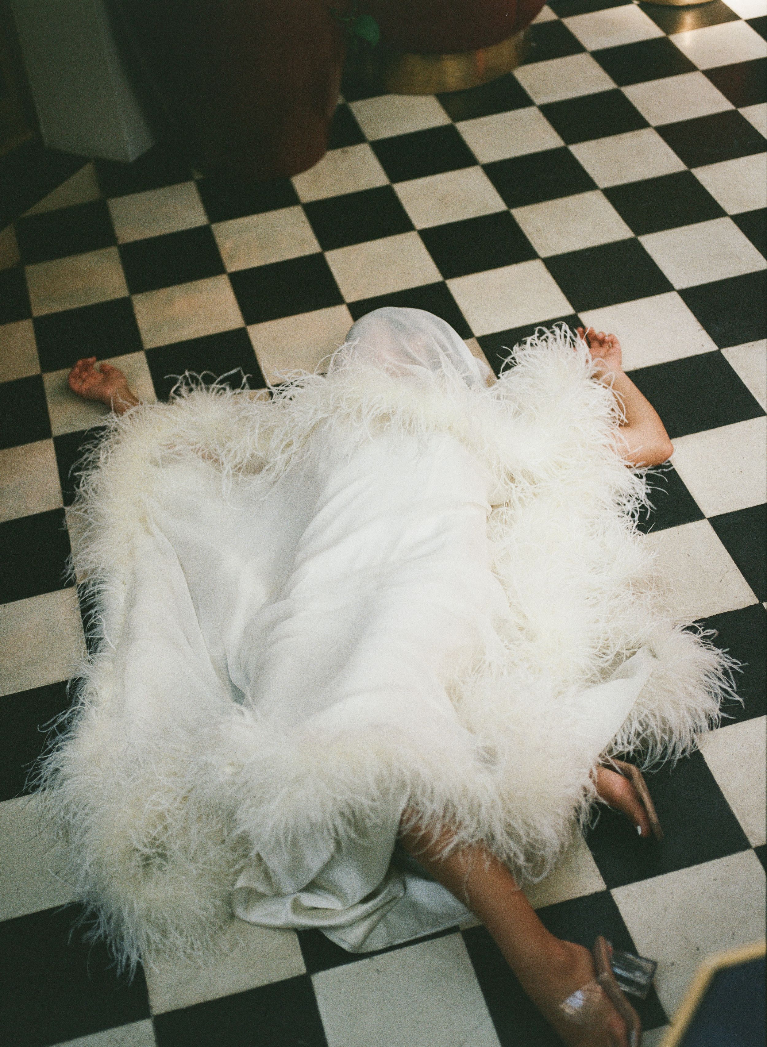 Woman in white feathered gown lying on black and white checkered floor, Paris bridal fashion editorial. Iconic, bold paris bride, film