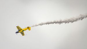 Yellow and green aerobatic plane performs a dramatic maneuver leaving a white smoke trail against a gray sky.