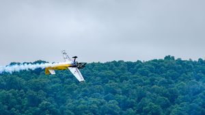 Yellow aerobatic plane performing a banking maneuver with smoke trail against a cloudy sky over lush green forest.