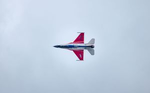 A red, white, and blue military fighter jet performing an aerial maneuver against a cloudy gray sky.