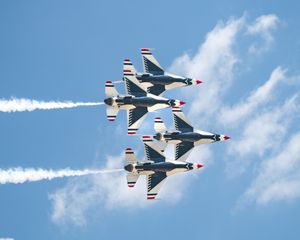 Four US Air Force Thunderbirds jets fly in tight diamond formation, trailing white smoke against a blue sky.