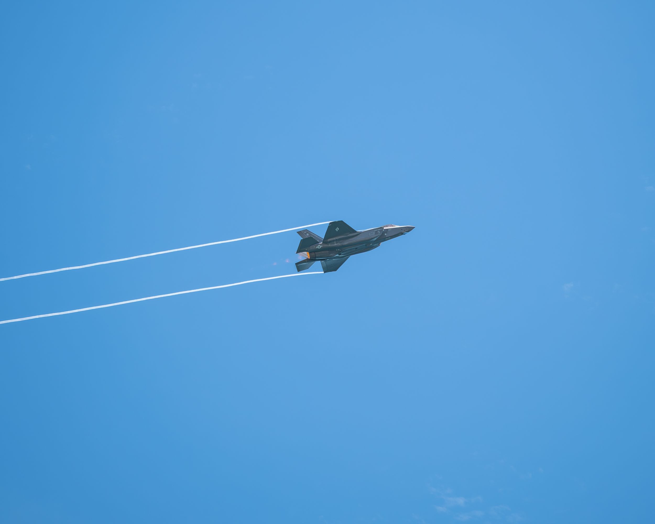 A military fighter jet soars through a clear blue sky leaving two white contrails in its wake.