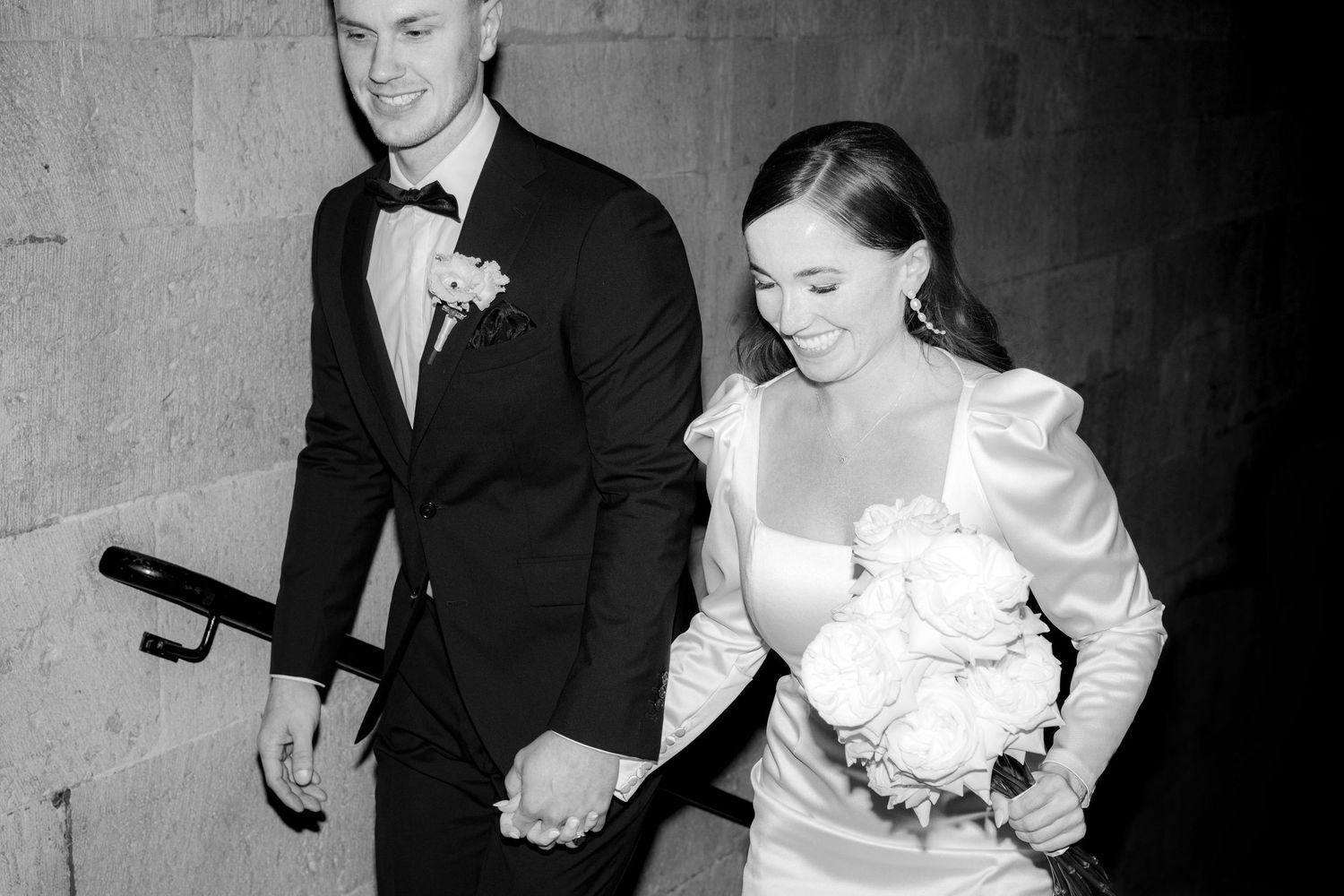 Black and white photo of a bride holding white roses bouquet and groom in tuxedo holding hands at wedding.