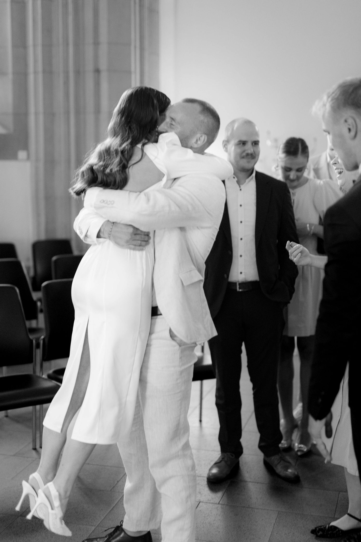 Black and white photo of a couple sharing a kiss and embrace at a wedding ceremony with guests in background.