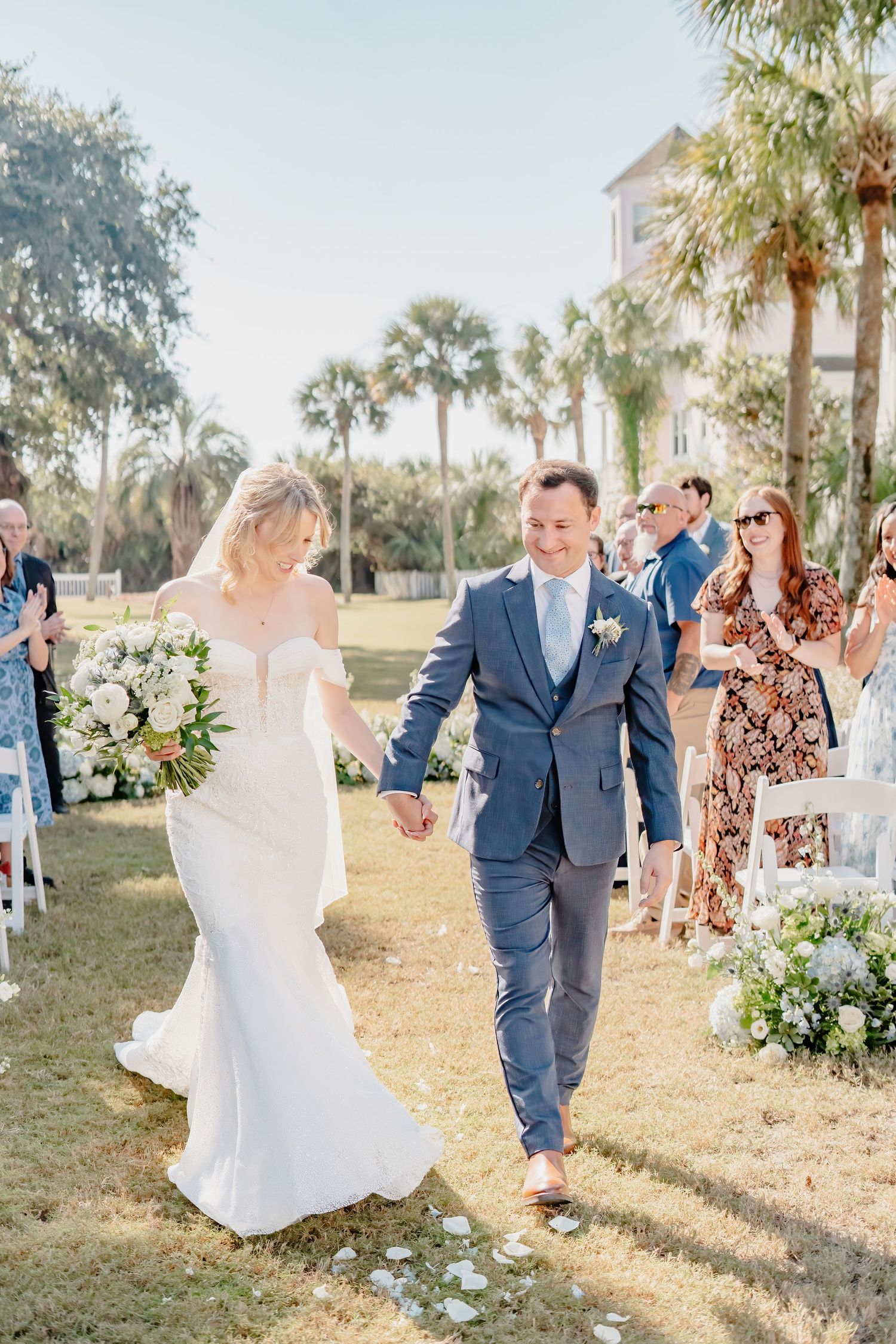 Bride and groom walk down outdoor aisle holding hands after wedding ceremony, surrounded by guests and palm trees.