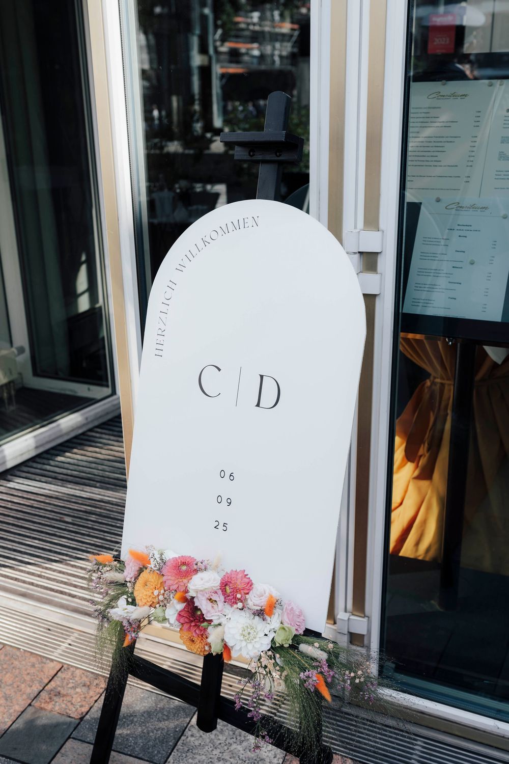 White wedding welcome sign with monogram C|D and date 08.09.25, adorned with pink and orange flowers on an easel.