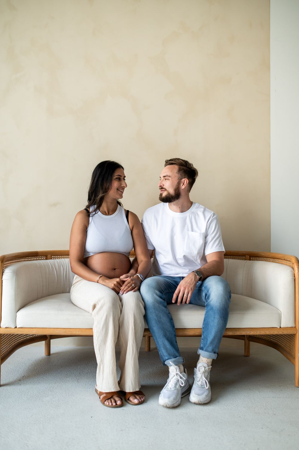 Pregnant couple sitting on a rattan sofa, smiling at each other, woman in white crop top showing baby bump.