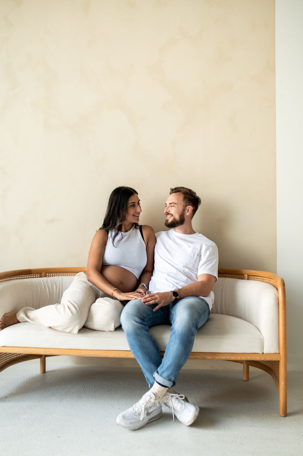 Expecting couple sitting on a wooden sofa, smiling at each other, woman in white top showing baby bump.