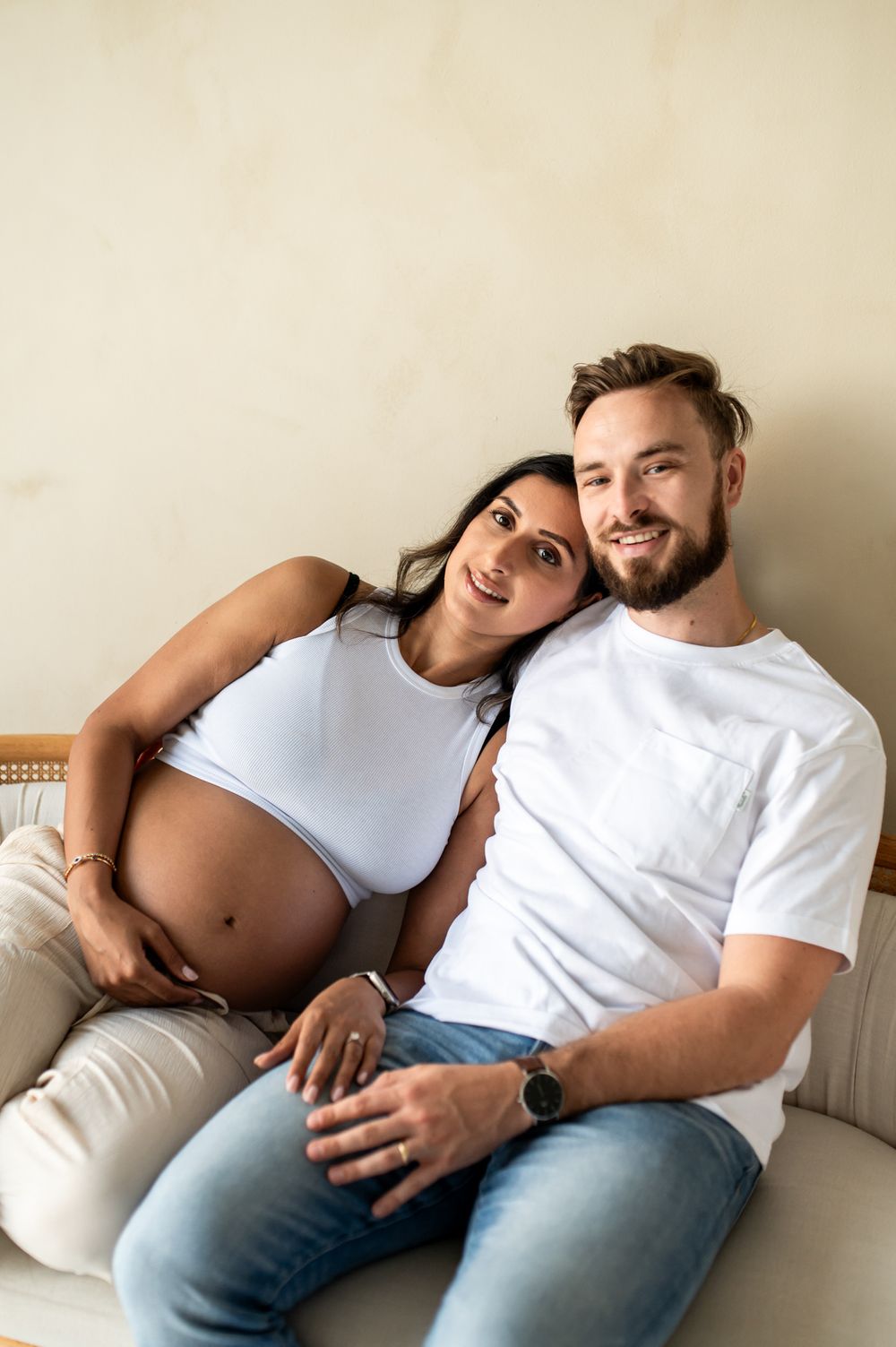Happy couple sitting on sofa, pregnant woman in white crop top leaning on smiling bearded man in white t-shirt.