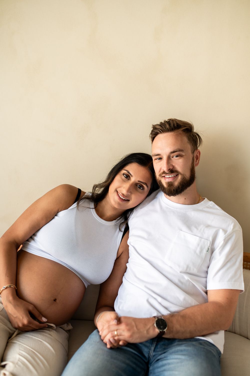 Smiling pregnant couple sitting together on a couch, woman leaning on mans shoulder, both in white tops.