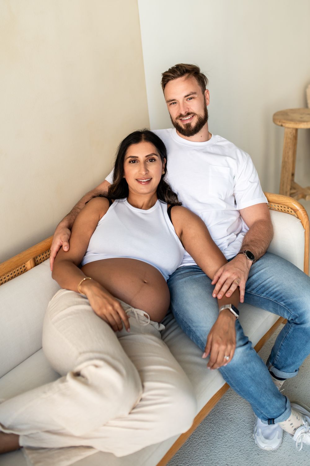 Pregnant woman in white crop top sits with partner on sofa, both smiling at camera during maternity photoshoot.