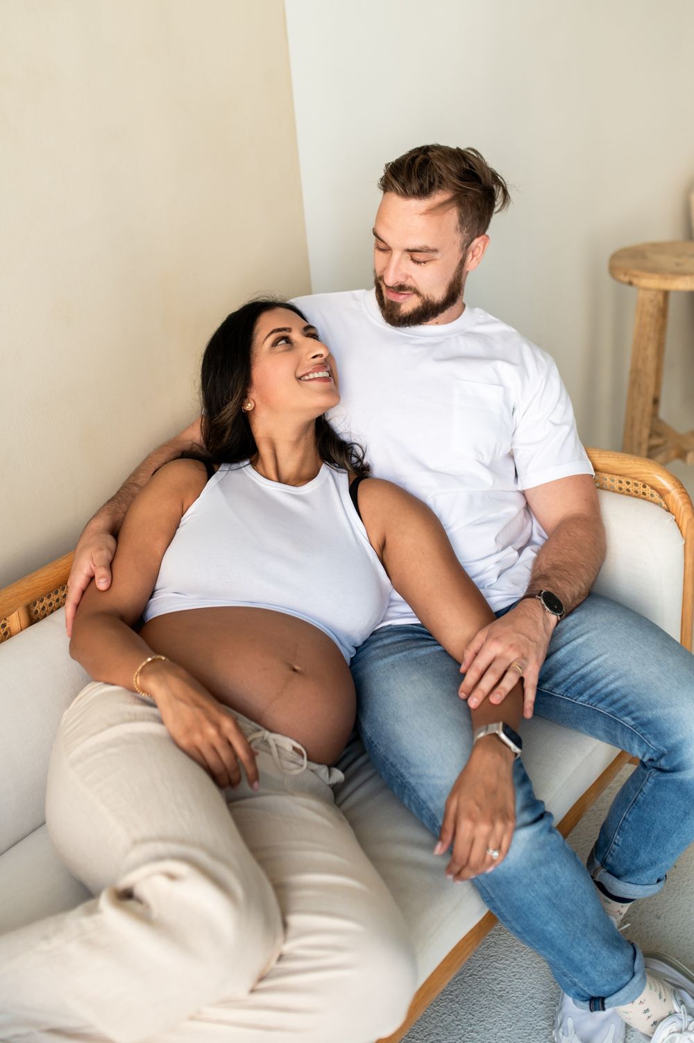 A pregnant woman smiles at her partner while they sit together on a couch, both wearing white tops and jeans.