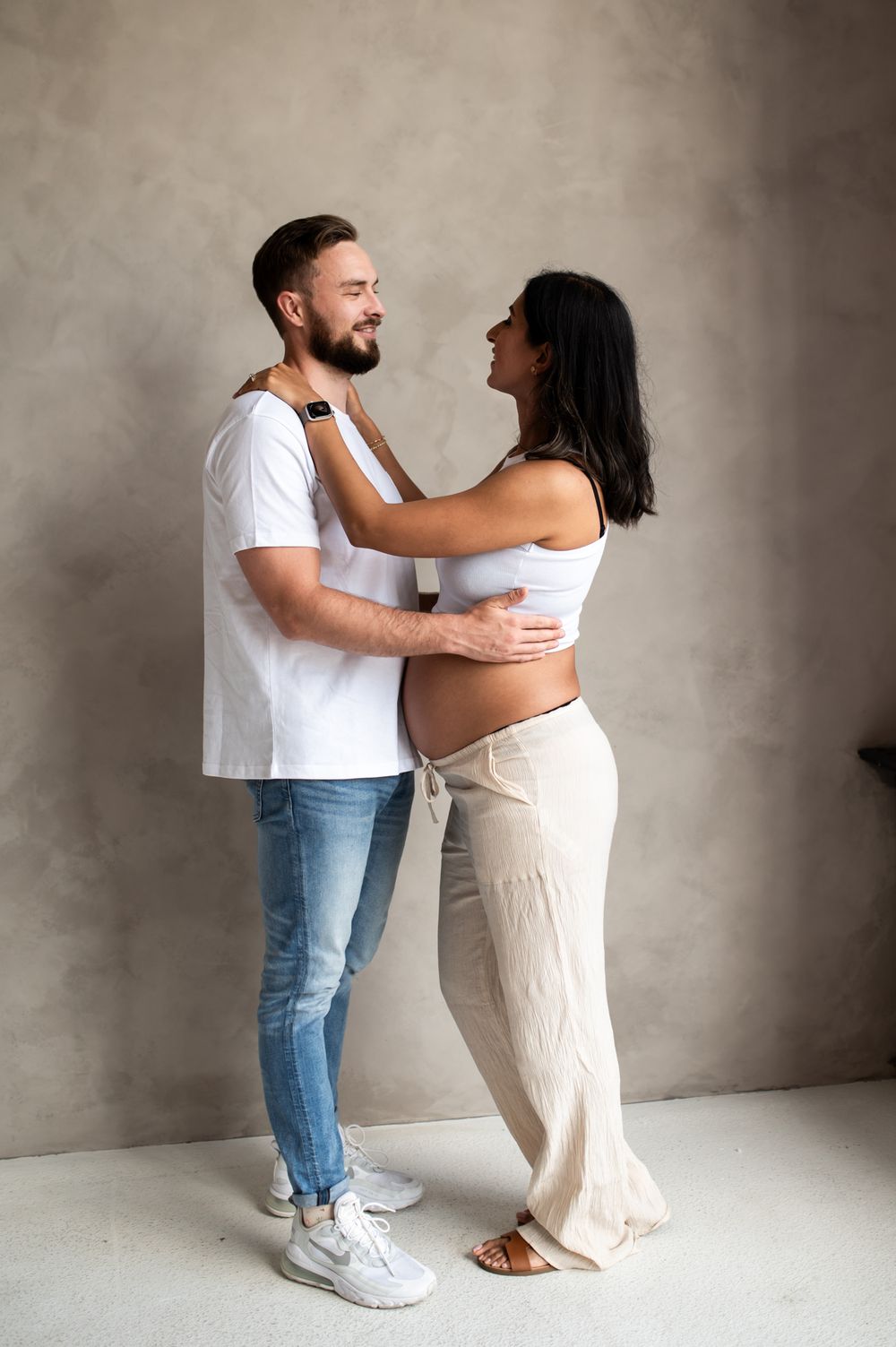 A happy couple shares a tender moment, with the pregnant woman in white crop top and beige pants embracing her smiling partner.