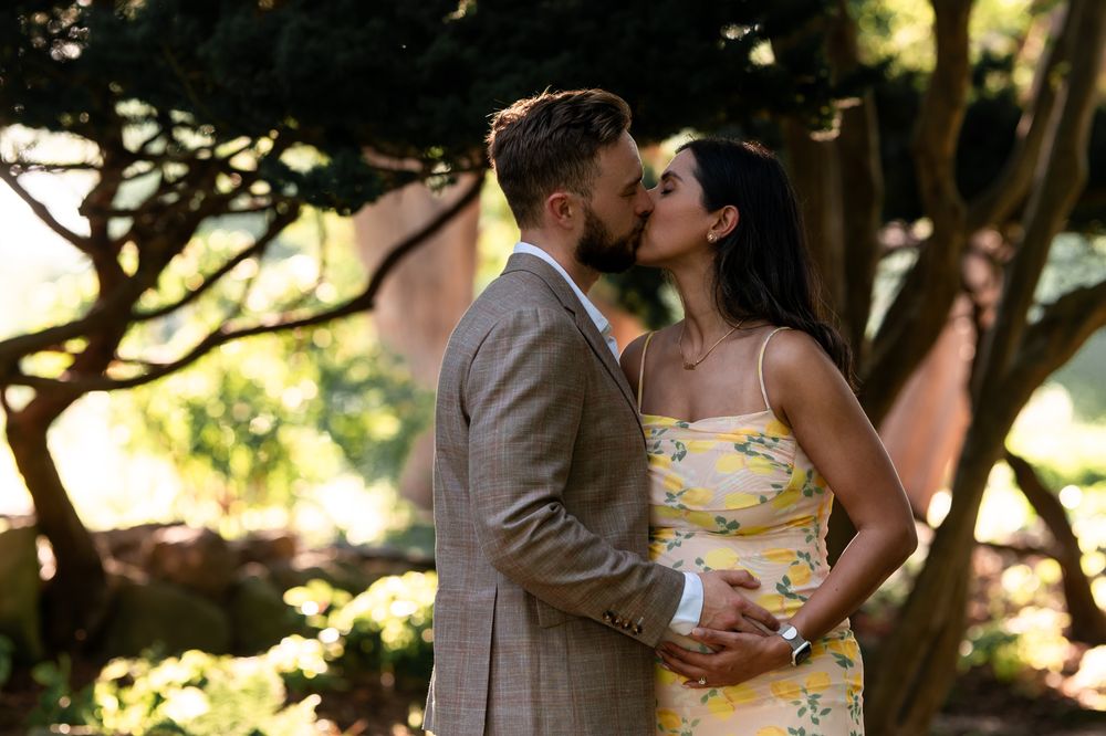 Couple sharing a romantic kiss outdoors, woman in yellow floral dress, man in gray suit, surrounded by lush green trees.