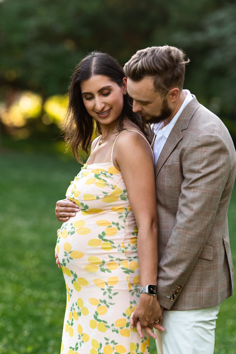 Couple maternity photo outdoors, woman in yellow lemon dress cradling baby bump, man in tan blazer embracing her.
