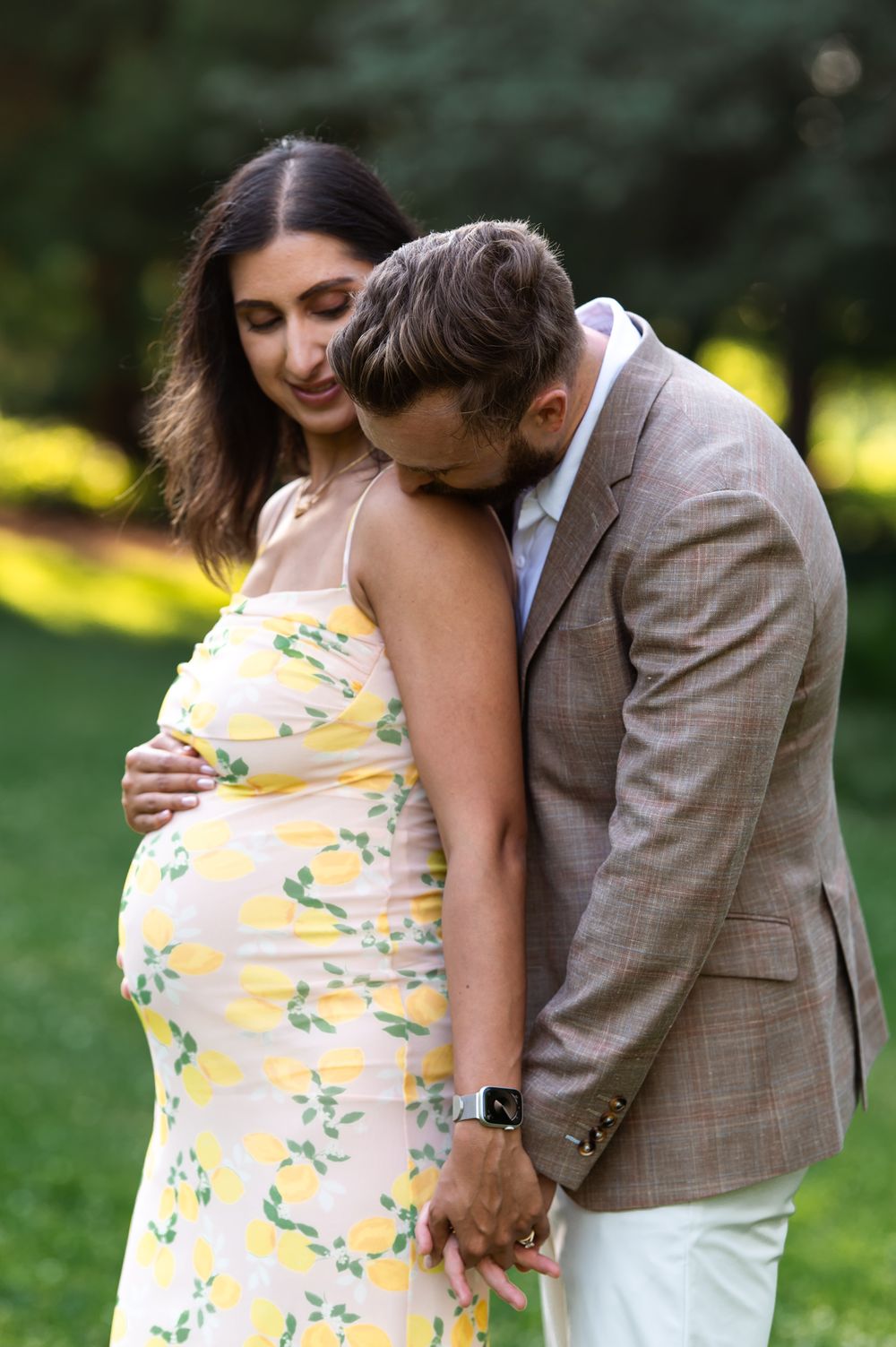 Pregnant woman in yellow floral dress with partner in tan suit sharing tender moment outdoors in lush green garden.