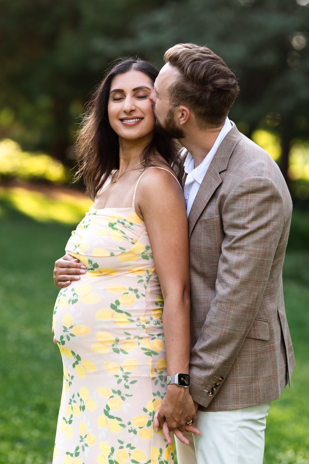 Pregnant woman in yellow floral dress smiles as man in brown blazer kisses her cheek outdoors.