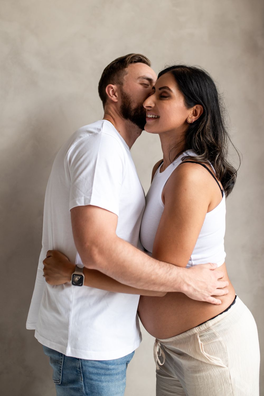 Expecting couple embracing and kissing during maternity shoot, both wearing white tops against gray background.
