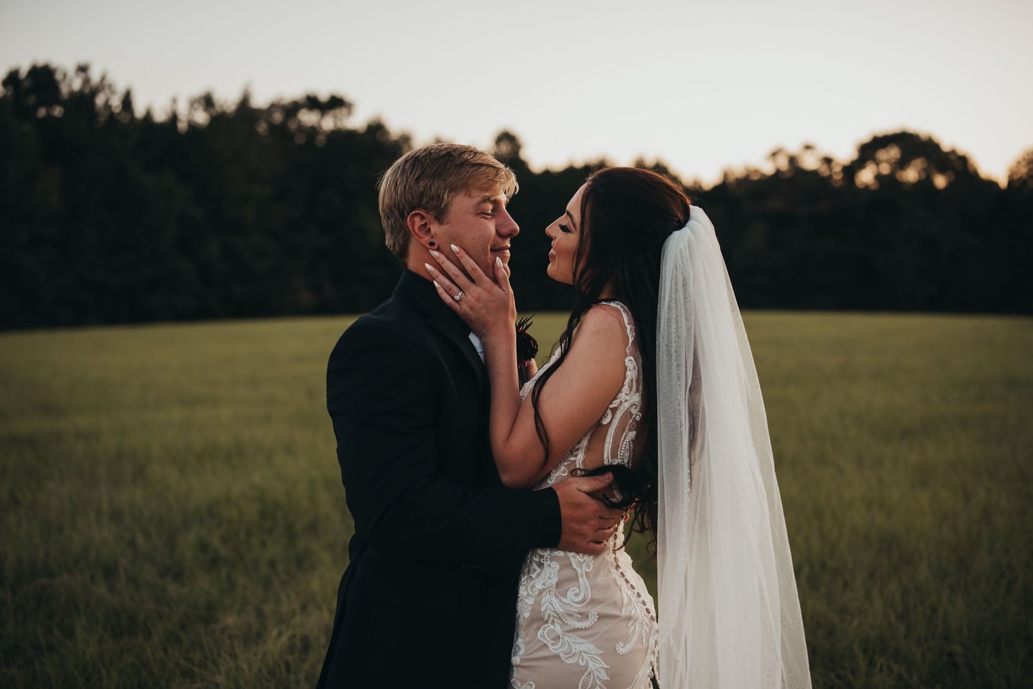 Bride and groom share a romantic moment at sunset in a lush green field, with the bride holding his face gently.